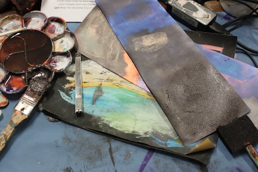 A collection of artistic tools and materials are spread across a surface, including a paint palette with various colors, brushes, and pieces of paper or canvas that feature abstract designs. The tools and materials are arranged in a casual, somewhat disorganized manner.