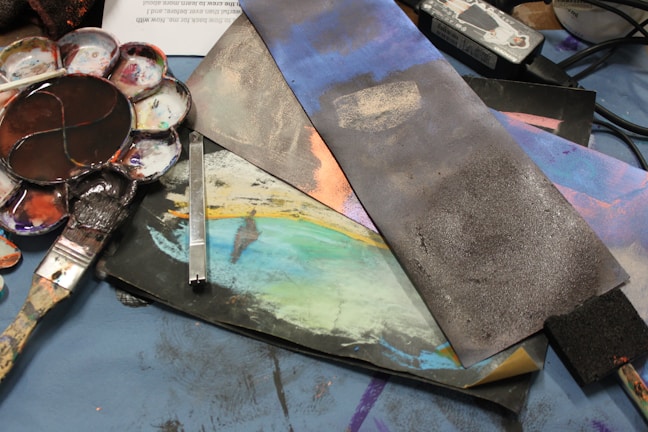 A collection of artistic tools and materials are spread across a surface, including a paint palette with various colors, brushes, and pieces of paper or canvas that feature abstract designs. The tools and materials are arranged in a casual, somewhat disorganized manner.