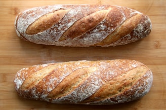 Two freshly baked baguettes with a golden brown crust rest on a wooden surface. The bread features distinct slits on top and is lightly dusted with flour, highlighting the artisanal texture.