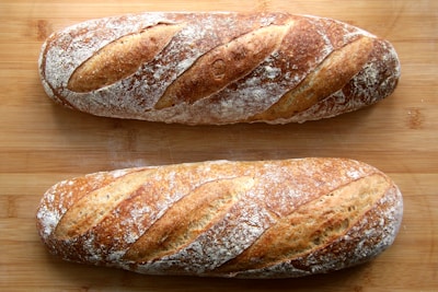 Two freshly baked baguettes with a golden brown crust rest on a wooden surface. The bread features distinct slits on top and is lightly dusted with flour, highlighting the artisanal texture.