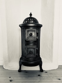 A vintage cast iron stove with ornate detailing, featuring a domed top and two small doors with engraved patterns. It stands against a textured white wall on a wooden floor.