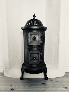 A vintage cast iron stove with ornate detailing, featuring a domed top and two small doors with engraved patterns. It stands against a textured white wall on a wooden floor.