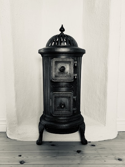 A vintage cast iron stove with ornate detailing, featuring a domed top and two small doors with engraved patterns. It stands against a textured white wall on a wooden floor.