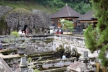 An ancient stone temple with intricate carvings surrounded by lush greenery. Visitors explore the site, with some near an open, sculpted cave entrance. Statues stand near a water feature, with the scene framed by trees and traditional thatched-roof structures.