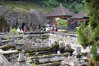 An ancient stone temple with intricate carvings surrounded by lush greenery. Visitors explore the site, with some near an open, sculpted cave entrance. Statues stand near a water feature, with the scene framed by trees and traditional thatched-roof structures.