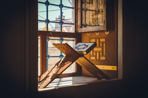 A Quran is placed on an intricately carved wooden stand positioned near a window. The sunlight illuminates the book, creating a warm and serene atmosphere. The background features a wooden panel with geometric patterns and a window with decorative latticework.