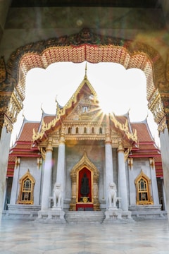 An ornate temple with intricate golden carvings, large pillars, and symmetrical architecture. Sunlight shines through the temple archway, highlighting a statue at the entrance. Two lion statues flank the steps leading up to the entrance.