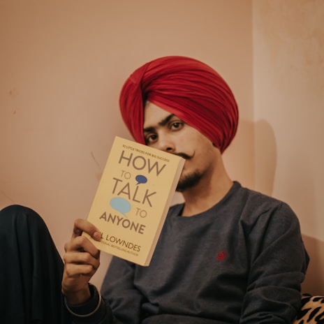 A person is sitting indoors, wearing a red turban and holding up a book titled 'How to Talk to Anyone'. The person has a mustache and is dressed in a dark sweater. The background features a neutral-colored wall, creating a calm setting.