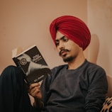 A person wearing a bright red turban is sitting and reading a book titled 'Fifty Shades of Grey' by E.L. James. The individual has a focused expression with a neatly groomed mustache, wearing a dark sweater, and is sitting against a muted cream-colored wall.