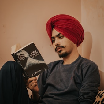 A person wearing a bright red turban is sitting and reading a book titled 'Fifty Shades of Grey' by E.L. James. The individual has a focused expression with a neatly groomed mustache, wearing a dark sweater, and is sitting against a muted cream-colored wall.