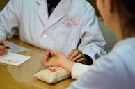 A person in a white coat appears to be taking the pulse of another person. They are seated at a wooden table with a notepad and a pen in view. The person receiving the pulse check rests their arm on a small pillow decorated with red and orange embroidery.