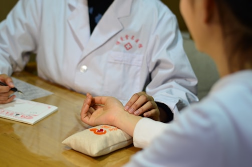A person in a white coat appears to be taking the pulse of another person. They are seated at a wooden table with a notepad and a pen in view. The person receiving the pulse check rests their arm on a small pillow decorated with red and orange embroidery.