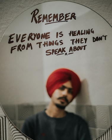 A reflective message is written on an oval mirror in bold, dark letters. The message reads: 'Remember everyone is healing from things they don't speak about.' In the background, a person wearing a red turban and dark clothing is faintly visible, reflected in the mirror but out of focus.