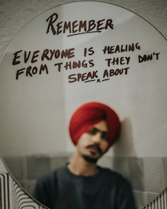 A reflective message is written on an oval mirror in bold, dark letters. The message reads: 'Remember everyone is healing from things they don't speak about.' In the background, a person wearing a red turban and dark clothing is faintly visible, reflected in the mirror but out of focus.