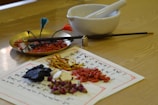 An assortment of traditional herbal ingredients, possibly used in Chinese medicine, arranged on papers with writing. A brass tray holds various plant materials next to a white mortar and pestle. A chopstick-style tool with colorful tassels is also present.