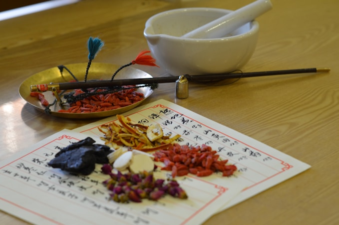 An assortment of traditional herbal ingredients, possibly used in Chinese medicine, arranged on papers with writing. A brass tray holds various plant materials next to a white mortar and pestle. A chopstick-style tool with colorful tassels is also present.