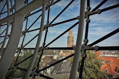 A modern, geometric steel framework creates a contrast with a historic stone tower visible in the background. The framework has black metal supports with circular joints, partially obscuring a view of rooftops featuring a mix of orange and gray tiles, and a few leafy trees.