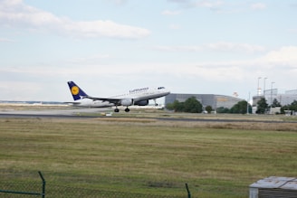 A commercial airplane in the process of landing on a runway. The aircraft is branded with the Lufthansa logo, featuring a white fuselage and a blue tail fin with a yellow logo. The surrounding area includes a grassy field in the foreground and airport buildings and infrastructure in the background. The sky is cloudy with patches of blue.