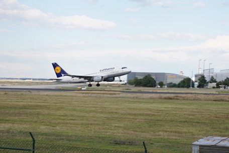 A commercial airplane in the process of landing on a runway. The aircraft is branded with the Lufthansa logo, featuring a white fuselage and a blue tail fin with a yellow logo. The surrounding area includes a grassy field in the foreground and airport buildings and infrastructure in the background. The sky is cloudy with patches of blue.