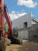 A construction site features a large orange excavator parked next to a partially constructed building. The building is made of concrete blocks and has scaffolding attached. The sky is clear with a few scattered clouds, and potted plants can be partially seen in the background.
