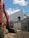 A construction site features a large orange excavator parked next to a partially constructed building. The building is made of concrete blocks and has scaffolding attached. The sky is clear with a few scattered clouds, and potted plants can be partially seen in the background.