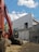 A construction site features a large orange excavator parked next to a partially constructed building. The building is made of concrete blocks and has scaffolding attached. The sky is clear with a few scattered clouds, and potted plants can be partially seen in the background.