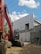 A construction site features a large orange excavator parked next to a partially constructed building. The building is made of concrete blocks and has scaffolding attached. The sky is clear with a few scattered clouds, and potted plants can be partially seen in the background.