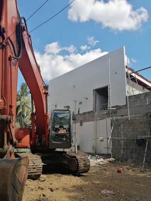 A construction site features a large orange excavator parked next to a partially constructed building. The building is made of concrete blocks and has scaffolding attached. The sky is clear with a few scattered clouds, and potted plants can be partially seen in the background.