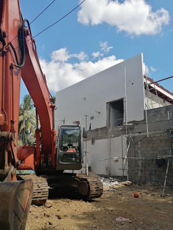 A construction site features a large orange excavator parked next to a partially constructed building. The building is made of concrete blocks and has scaffolding attached. The sky is clear with a few scattered clouds, and potted plants can be partially seen in the background.