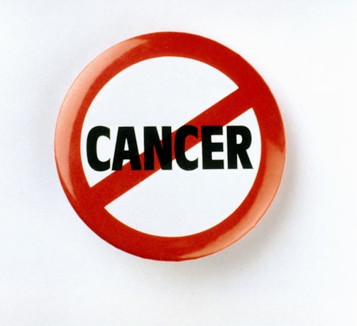 A circular badge with a red border and slash through the word 'CANCER' printed in bold black letters on a white background, symbolizing opposition or prohibition.