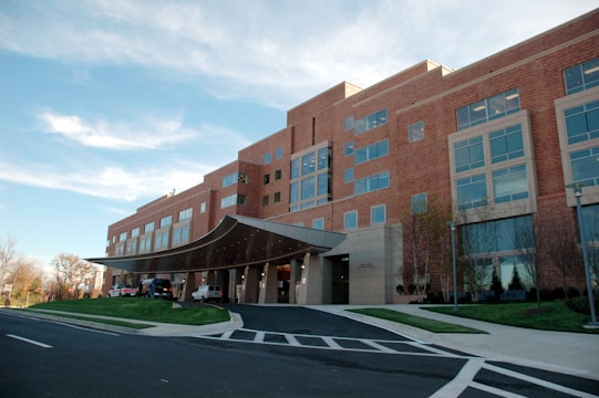 A modern multi-story brick building with large windows and a prominent entrance canopy. The structure is surrounded by neatly manicured lawns and a clear roadway. The sky overhead is clear with a few clouds.