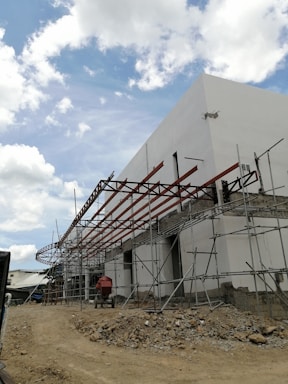 A construction site with a partially built structure featuring scaffolding and a framework for a roof. The ground is covered in dirt and rubble, and there is construction equipment present. The sky is partly cloudy, creating a bright and open atmosphere.