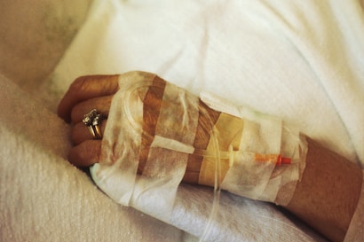 A close-up of an elderly person's hand with a medical IV line inserted, secured with medical tape. The hand is resting on a soft white fabric, and the person is wearing a gold ring with a gemstone.