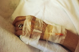 A close-up of an elderly person's hand with a medical IV line inserted, secured with medical tape. The hand is resting on a soft white fabric, and the person is wearing a gold ring with a gemstone.