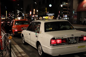 A nighttime urban street scene with several taxis lined up along the road. A white taxi and an orange taxi are prominently visible. Bright storefront signs, including a Starbucks, are illuminated in the background. Pedestrians are walking on the sidewalk, and the atmosphere is lively with city lights and signage.