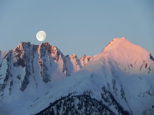 A snow-plastered mountain peak glowing pink at alpenglow with a pale moon above
