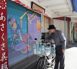 An elderly man wearing a black hat and striped shirt stands next to a bicycle with a basket full of cleaning supplies. He appears to be cleaning or painting a closed shop shutter that features vibrant artwork, depicting a traditional Japanese figure. The scene is illuminated by daylight, and there are additional closed shutters and a red signboard with Japanese text in the background.