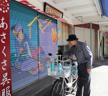 An elderly man wearing a black hat and striped shirt stands next to a bicycle with a basket full of cleaning supplies. He appears to be cleaning or painting a closed shop shutter that features vibrant artwork, depicting a traditional Japanese figure. The scene is illuminated by daylight, and there are additional closed shutters and a red signboard with Japanese text in the background.