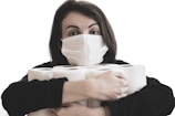 A person wearing a medical face mask is holding several rolls of toilet paper. The individual has dark hair and gazes directly at the camera. The background is plain and light-colored, highlighting the focus on the person and the items they are holding.