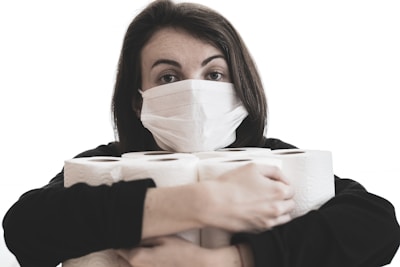 A person wearing a medical face mask is holding several rolls of toilet paper. The individual has dark hair and gazes directly at the camera. The background is plain and light-colored, highlighting the focus on the person and the items they are holding.