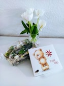 A small bouquet of white tulips in a glass vase is placed on a white surface against a textured white wall. Next to it, there is a decorative arrangement with greenery and small eggs wrapped in clear plastic. A square gift box with an illustration of a plush bear holding a red flower, accompanied by the text 'For You', is positioned on the right.