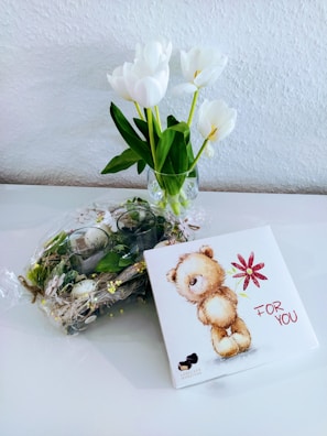 A small bouquet of white tulips in a glass vase is placed on a white surface against a textured white wall. Next to it, there is a decorative arrangement with greenery and small eggs wrapped in clear plastic. A square gift box with an illustration of a plush bear holding a red flower, accompanied by the text 'For You', is positioned on the right.
