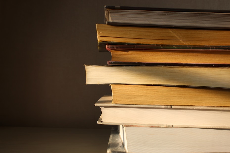 A stack of books with worn, textured covers and pages in warm lighting, creating a slightly nostalgic atmosphere. The books are arranged horizontally, with shadowed creases and folds visible on the edges.