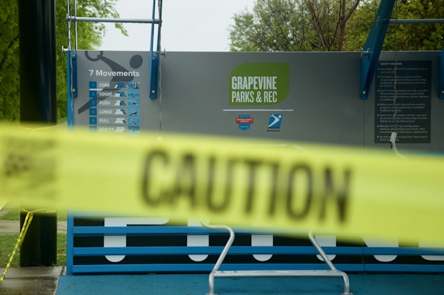 A caution tape with bold black letters spans across the foreground, partially obscuring a public workout area behind it. The words 'Grapevine Parks & Rec' are visible on a signboard, along with a list of seven movements like core, squat, push, and more. The area appears to be closed off for safety reasons.