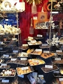 A display window filled with a variety of chocolates and confections, neatly arranged in trays with labels showing names and prices. The background features decorative items such as champagne bottles and porcelain figurines. The setting is elegant, with a focus on luxury chocolate products.