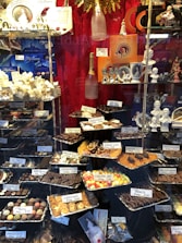 A display window filled with a variety of chocolates and confections, neatly arranged in trays with labels showing names and prices. The background features decorative items such as champagne bottles and porcelain figurines. The setting is elegant, with a focus on luxury chocolate products.