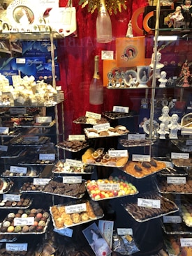 A display window filled with a variety of chocolates and confections, neatly arranged in trays with labels showing names and prices. The background features decorative items such as champagne bottles and porcelain figurines. The setting is elegant, with a focus on luxury chocolate products.