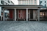 An outdoor theater entrance with a brick facade featuring large letters spelling out 'FAIR.' The entrance is supported by columns, and there are benches in front. Bicycles are parked on the left side and several windows with green shutters are visible on the adjacent buildings. A cobblestone street stretches in front of the theater.