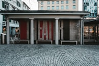 An outdoor theater entrance with a brick facade featuring large letters spelling out 'FAIR.' The entrance is supported by columns, and there are benches in front. Bicycles are parked on the left side and several windows with green shutters are visible on the adjacent buildings. A cobblestone street stretches in front of the theater.