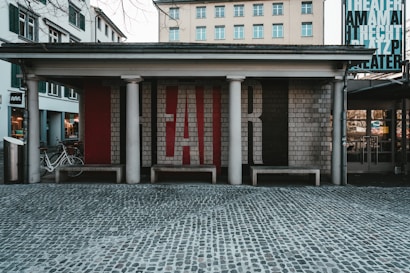 An outdoor theater entrance with a brick facade featuring large letters spelling out 'FAIR.' The entrance is supported by columns, and there are benches in front. Bicycles are parked on the left side and several windows with green shutters are visible on the adjacent buildings. A cobblestone street stretches in front of the theater.